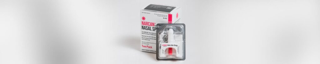 a photograph of a narcan nasal spray, still in sterile packaging, in front of the box it comes packaged in.