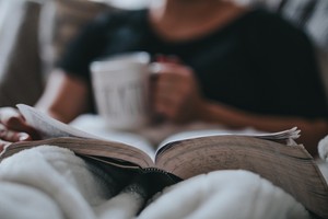 A slightly out of focus photo. It shows a person sitting on a couch or bed, they have a book open on their lap and a coffee mug in their hand.