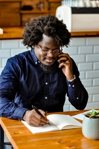 An African American man sits at a table in a coffee shop. He is writing in a notebook with his right hand while he holds a phone to his left ear with his left hand.
