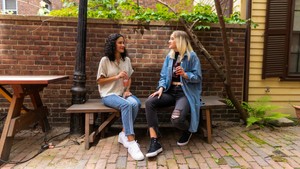 two women sit on a bench in an outdoors setting. The ground is pavers, there is a brick behind them. Behind the brick wall we can see some plants and more buildings. They women are having a pleasant conversation and enjoying soft drinks.