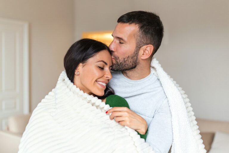 a photograph of a couple sitting together with a blanket around them, the man is kissing the woman on the forehead